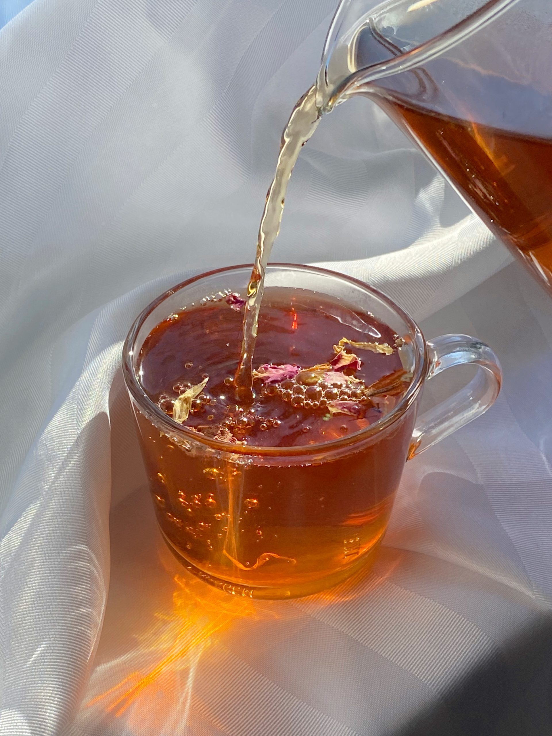 A rooibos loose leaf tea blend being pored delictely from a glass pitcher into a glass mug. Rose petals sit on top of the tea and small bubbles appear as the tea pours. The sun shines through the pouring tea to cast light and shadows on the surrounding soft white sheet. T. Kettle Loose Leaf Tea: Premium Quality, Environmental Responsibility, Social Equity.