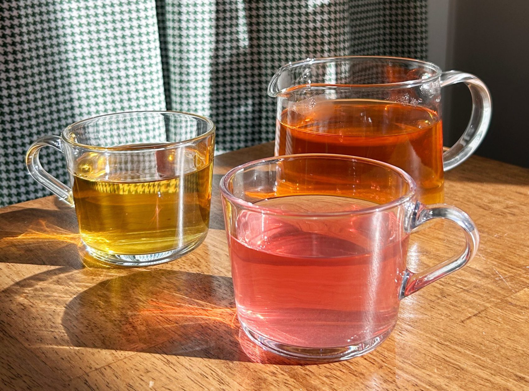 Colourful tea sits upon a wooden table in clear mugs. They are auburn, Golden, and Pink tea blends. Light shines through illuminating the vibrancy of the tea and casts light shadows. Teddy's Tea Club.