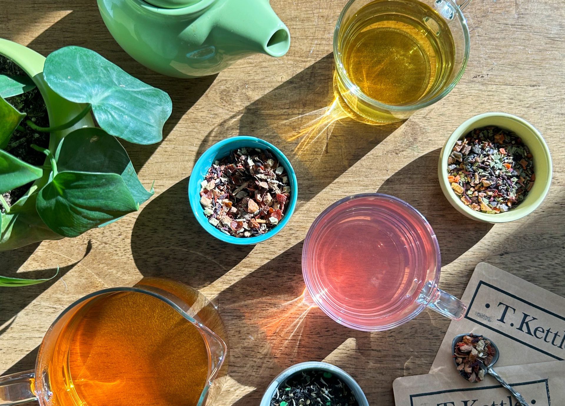 Overhead shot of colourful loose leaf tea in glass mugs, a small green teapot, and a variety of loose leaf tea blends distributed on a wooden table in colourful pinch pots for a curated tea aesthetic. Explore the world of premium loose leaf teas with T. Kettle.