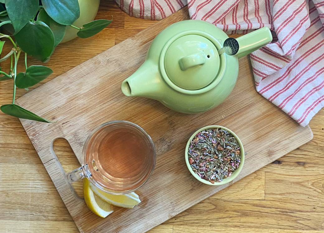Overhead shot of a green tea pot, raspberry lemon verbena loose leaf tea in a green pinch pot, a light pink tea in a glass mug, and two lemon slices. The assortment rests on a wooden cutting board, placed on top a wooden countertop. Dark green leaves of a house plant peek over the corners, and a tea towel is cast around the tea pot to frame the image. Experience premium loose leaf teas for wellness with T. Kettle.
